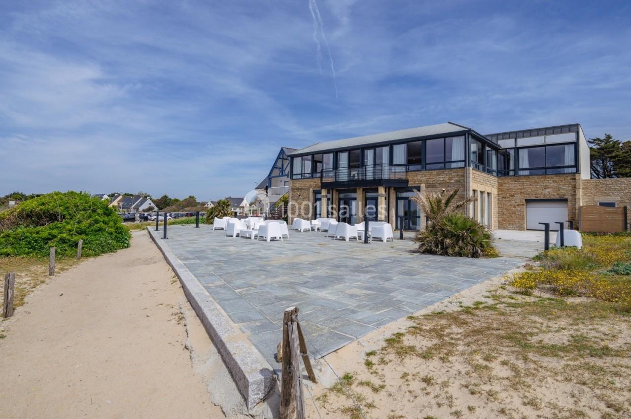 Bâtiment moderne en bord de mer avec terrasse pavée, mobilier extérieur blanc et végétation côtière.