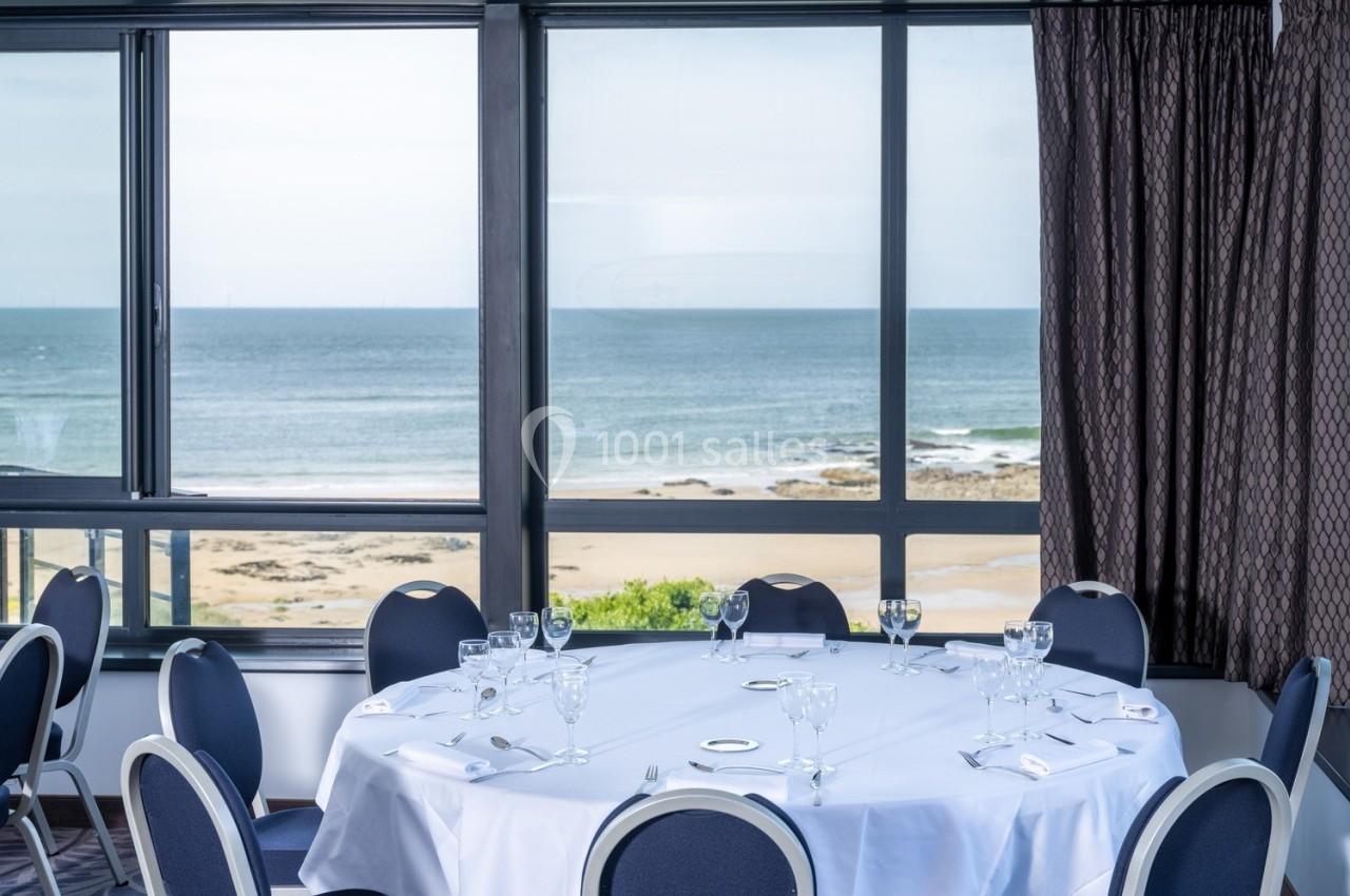 Salle de réunion avec table ronde dressée, vue sur la plage et l'océan à travers de grandes baies vitrées.
