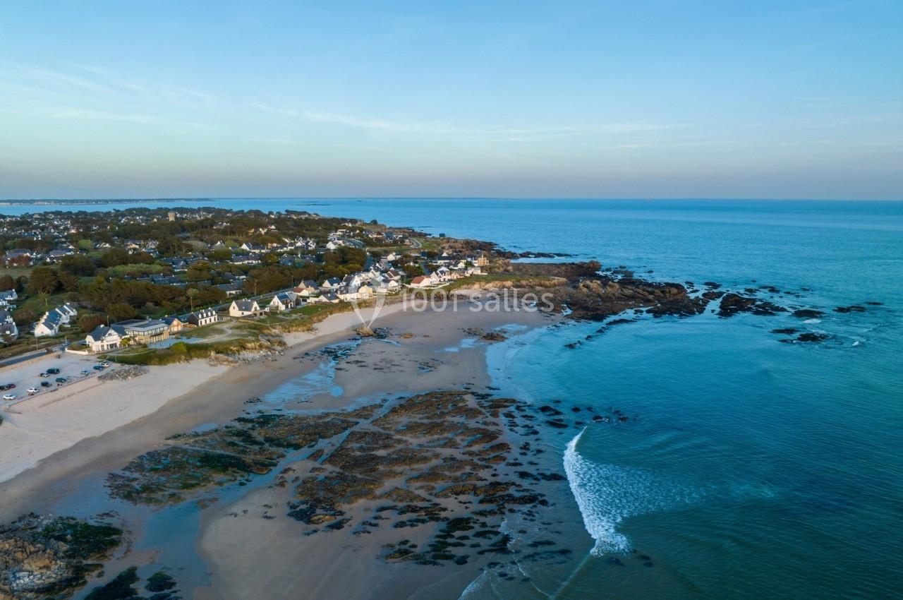 Vue aérienne d'une plage bordée de maisons, avec des rochers et l'océan s'étendant sous un ciel dégagé.