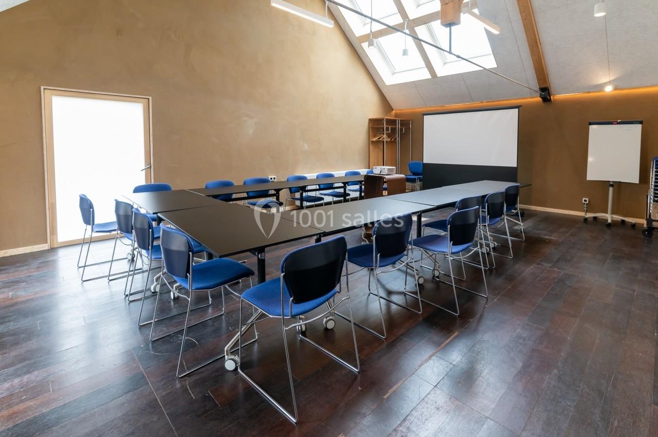 Salle de réunion lumineuse avec tables disposées en U, chaises bleues, tableau blanc et écran de projection.