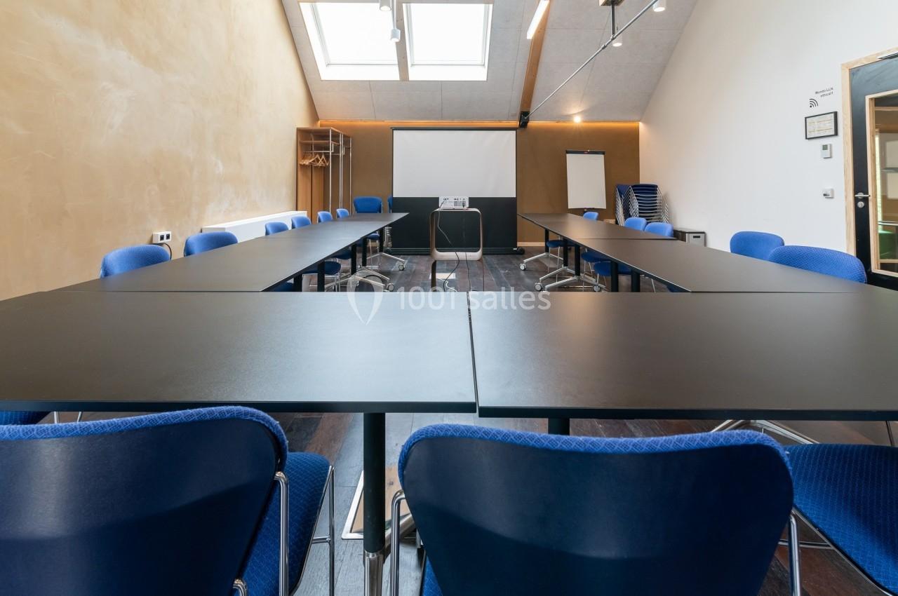 Salle de réunion lumineuse avec tables noires disposées en U, chaises bleues et écran de projection à l'arrière.
