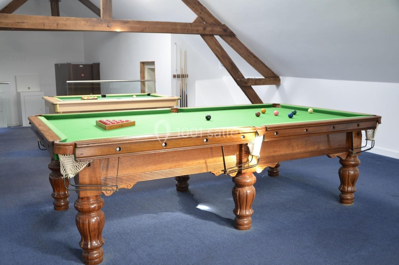 Deux tables de billard en bois avec tapis vert, situées dans une pièce lumineuse aux poutres apparentes.