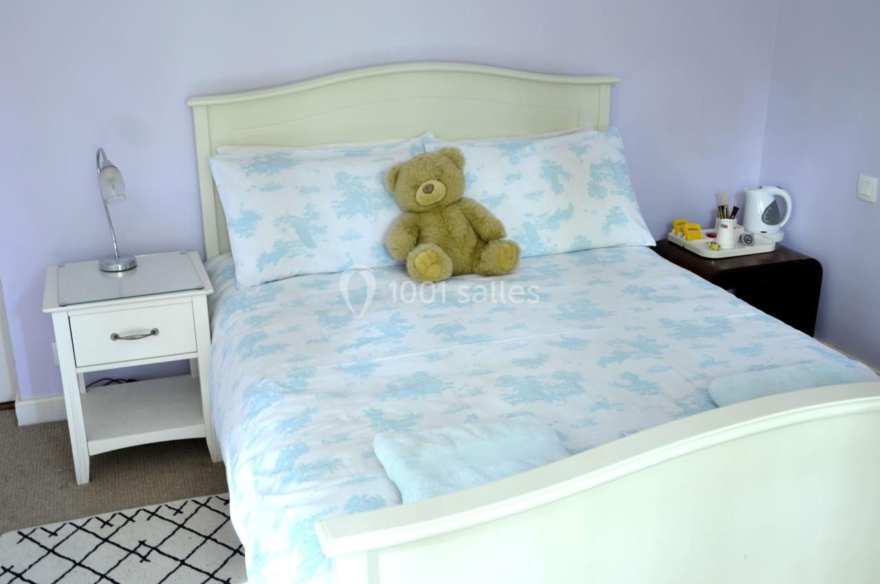 Lit double avec draps bleus à motifs, un ours en peluche posé dessus, entouré de tables de chevet avec lampe et accessoires.