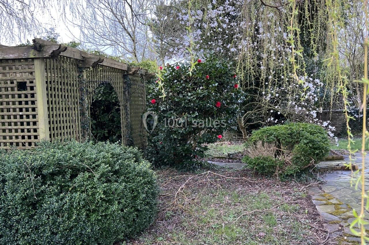 Jardin avec pergola en bois, arbustes taillés, fleurs rouges et blanches, et branches tombantes d'un saule.