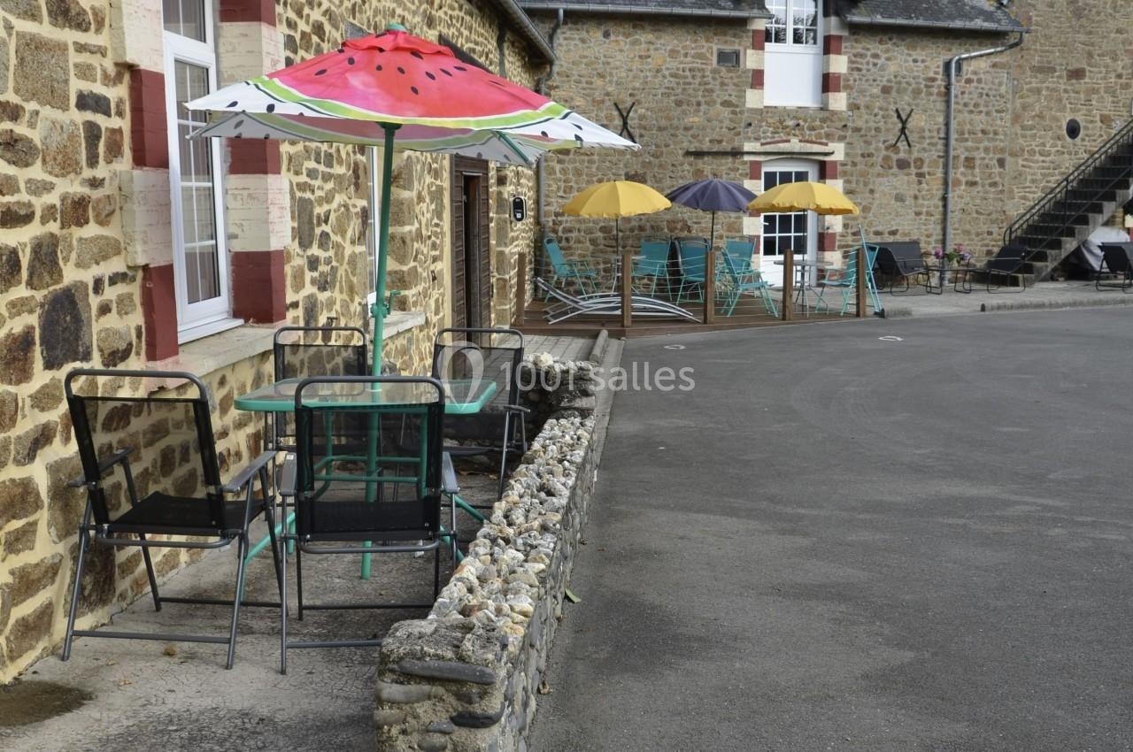 Table avec chaises noires sous un parasol coloré en forme de pastèque, près d'une maison en pierre et d'une terrasse.