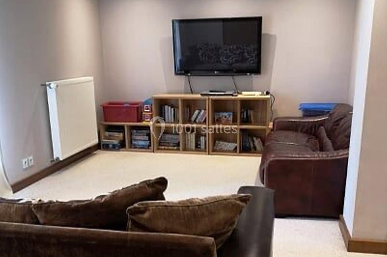 Salon avec canapé, fauteuil, étagères remplies de livres et objets, télévision murale et radiateur sur le mur.