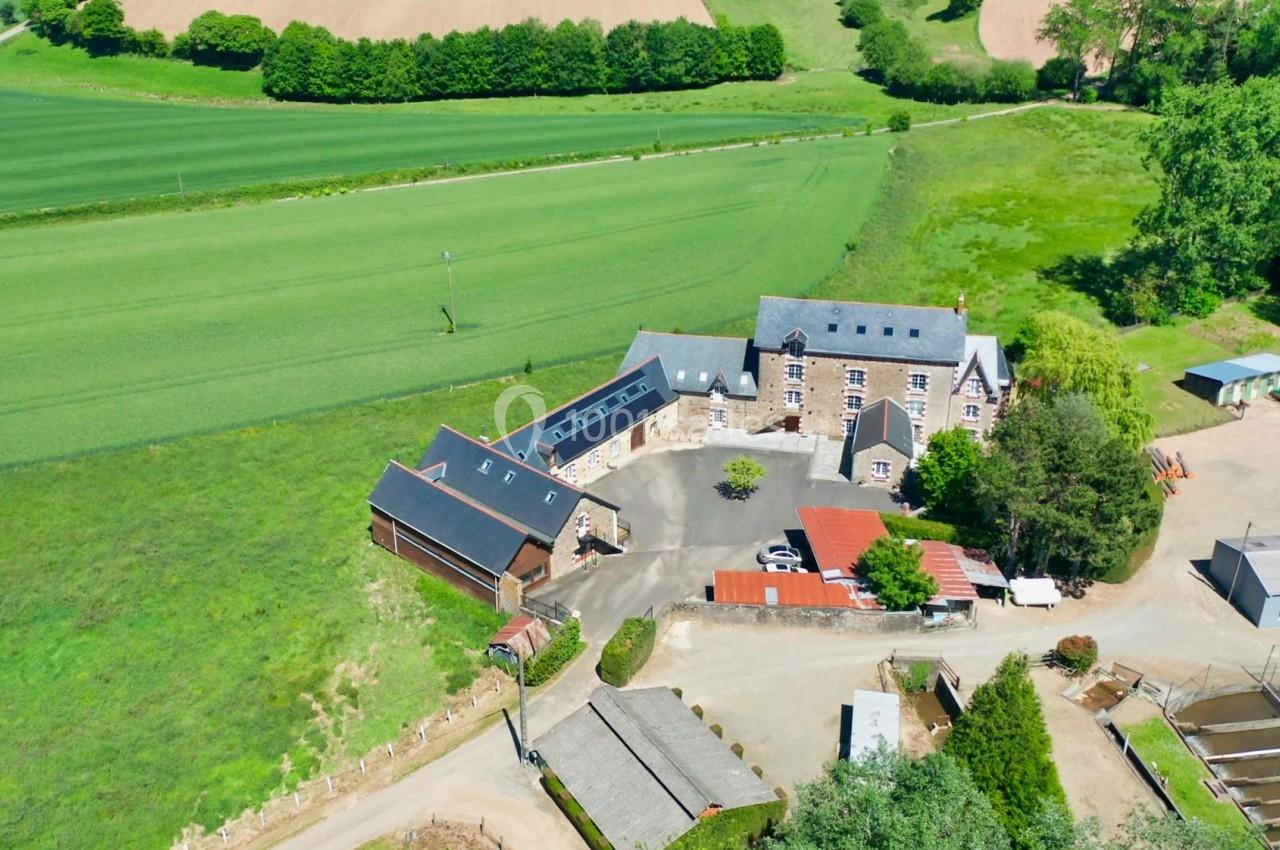 Vue aérienne d'une ferme avec bâtiments en pierre, toits en ardoise, champs verts et arbres environnants.