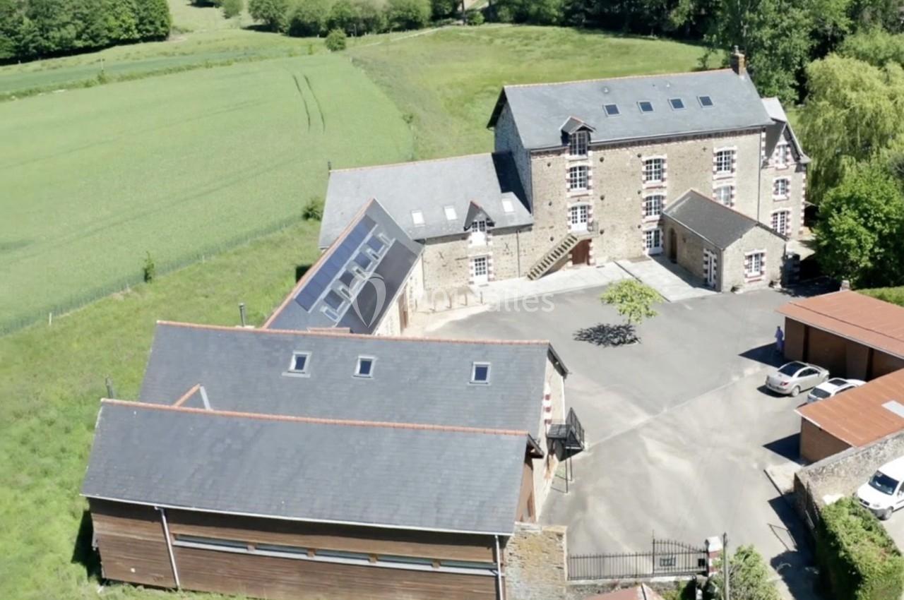 Vue aérienne d'un bâtiment en pierre avec dépendances modernes, entouré de champs et d'arbres.