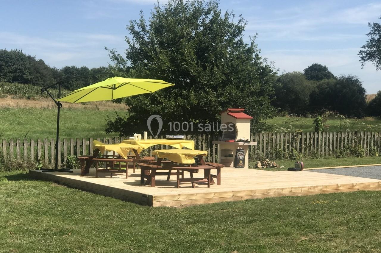 Terrasse en bois avec tables, chaises, parasol vert et barbecue, entourée de pelouse et d'arbres sous un ciel dégagé.