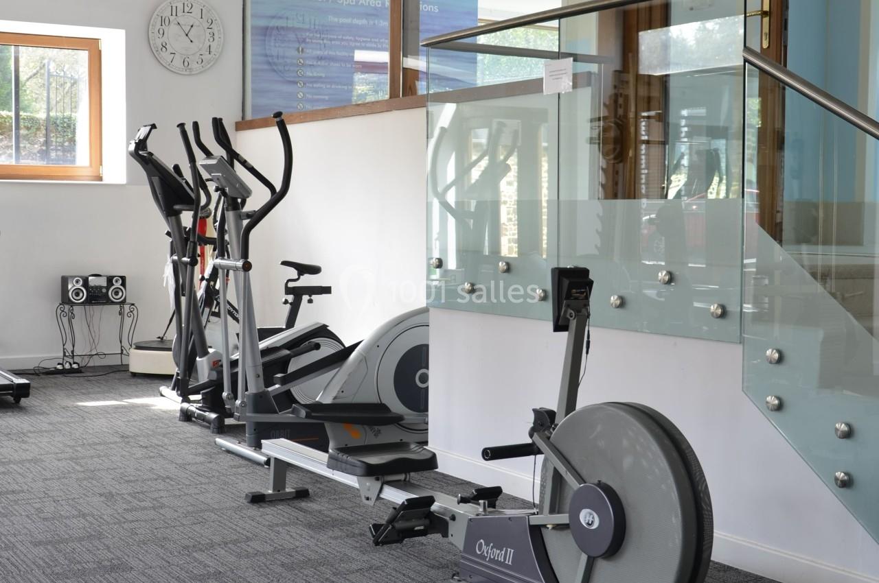 Salle de sport lumineuse avec vélos elliptiques, rameur et équipements de fitness près d'une rambarde en verre.