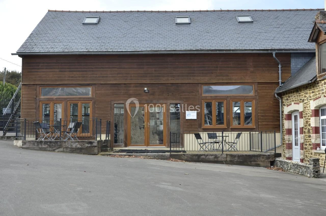 Façade d'un bâtiment en bois avec plusieurs fenêtres, une porte vitrée et une terrasse équipée de tables et chaises.