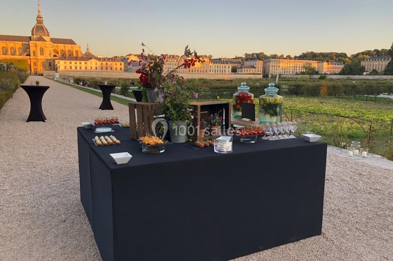 Table de buffet dressée en extérieur avec des amuse-bouches, des fleurs et des boissons, devant un paysage de jardin et…