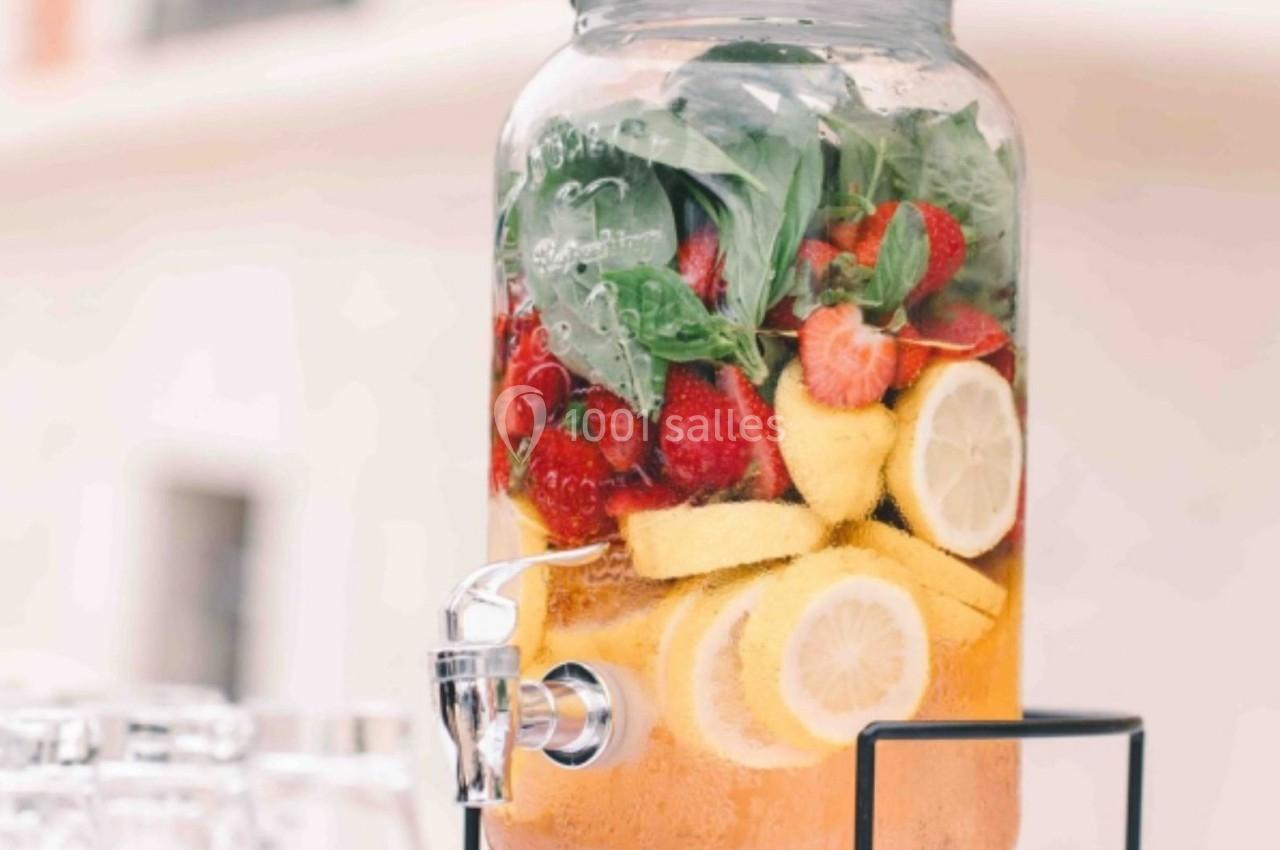 Distributeur d'eau infusée avec des fraises, des citrons, des framboises et des feuilles de menthe, posé sur un support noir.