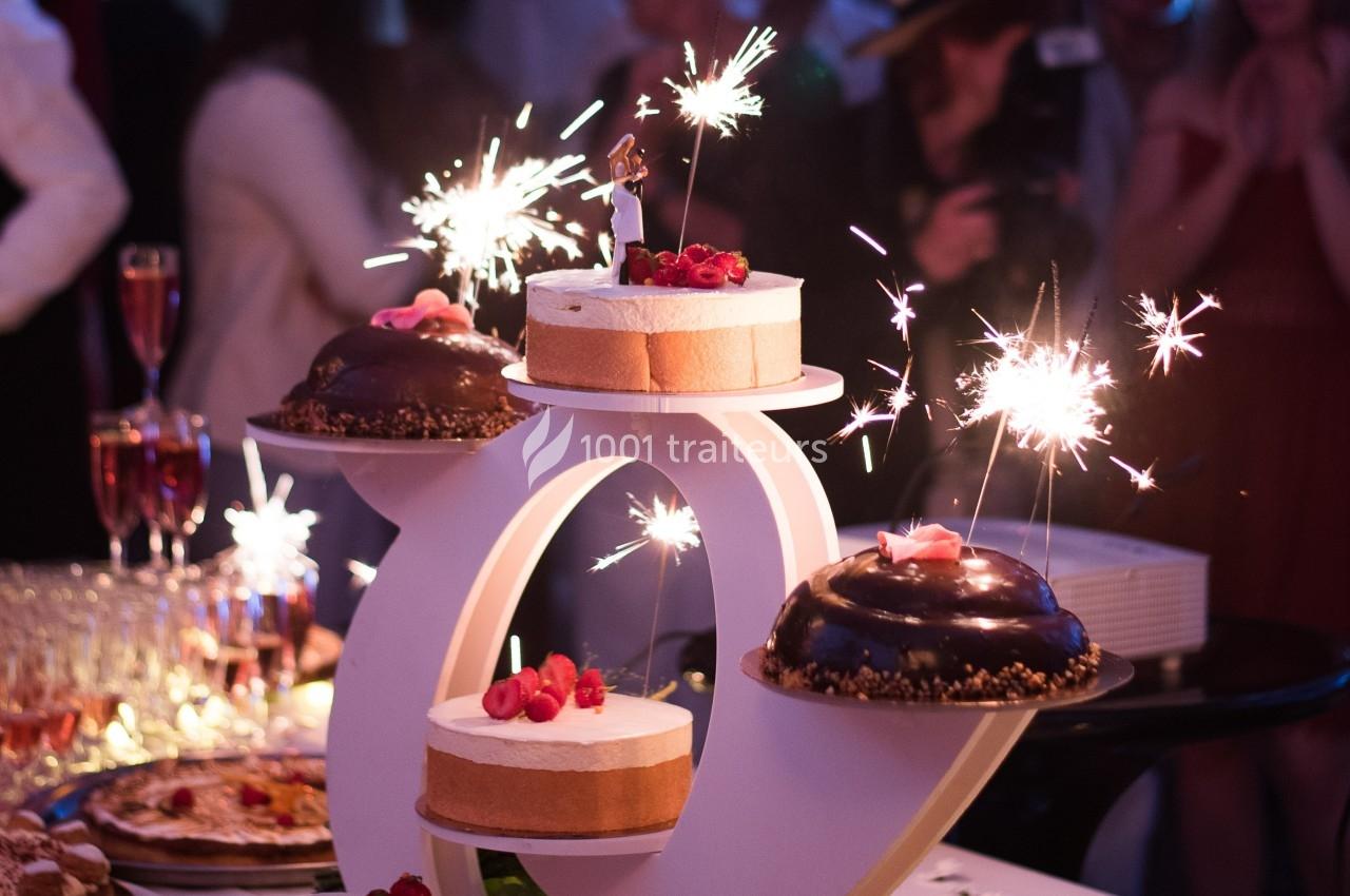 Gâteau de fête décoré de bougies scintillantes, entouré de desserts et de convives en arrière-plan flou.