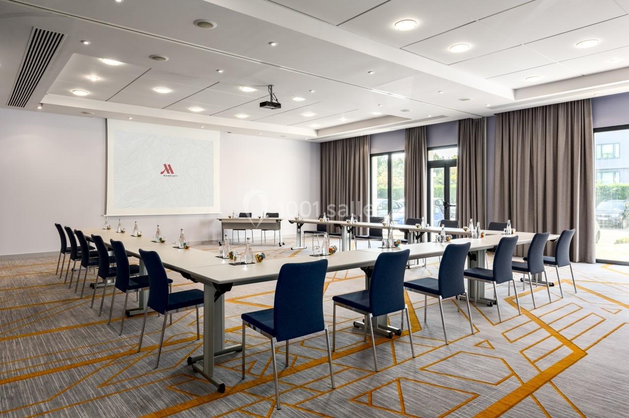 Salle de réunion moderne avec tables disposées en U, chaises bleues, écran de projection et lumière naturelle.