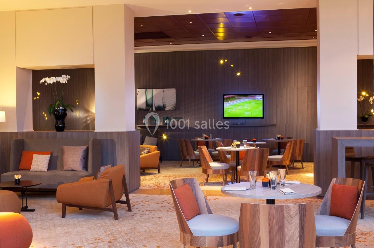 Espace lounge moderne avec fauteuils, tables basses, éclairage chaleureux et écran diffusant un match de sport.