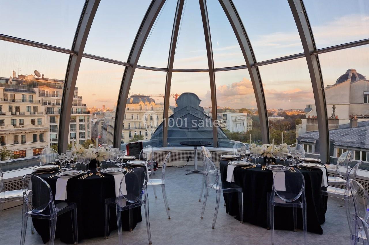 Salle à manger élégante sous une verrière offrant une vue panoramique sur des bâtiments parisiens au coucher du soleil.