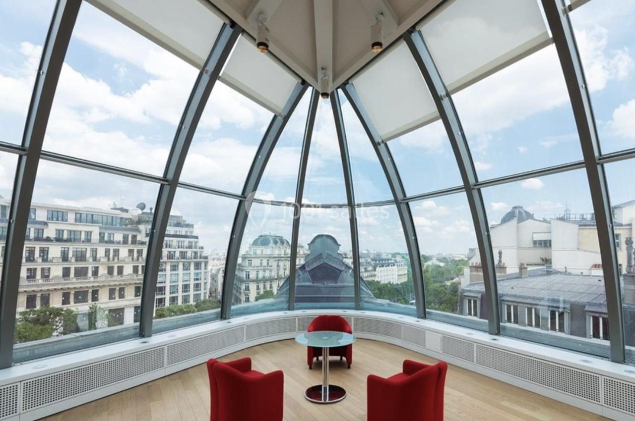 Salle lumineuse avec verrière arrondie, deux fauteuils rouges et une table en verre, vue sur des bâtiments urbains.