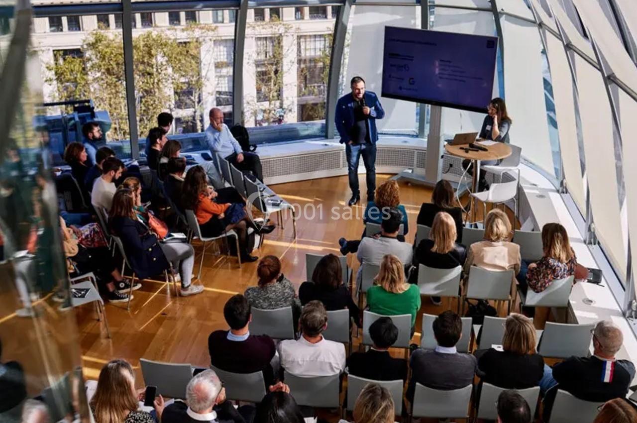 Un conférencier s'adresse à un public assis dans une salle lumineuse avec de grandes baies vitrées.
