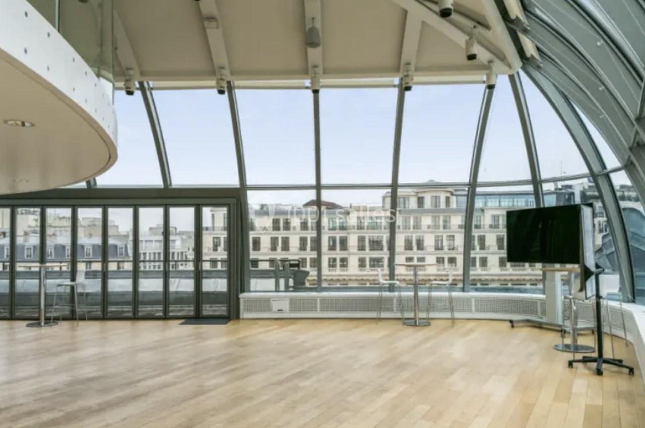 Salle lumineuse avec parquet en bois, grandes baies vitrées courbées et vue sur des bâtiments urbains.
