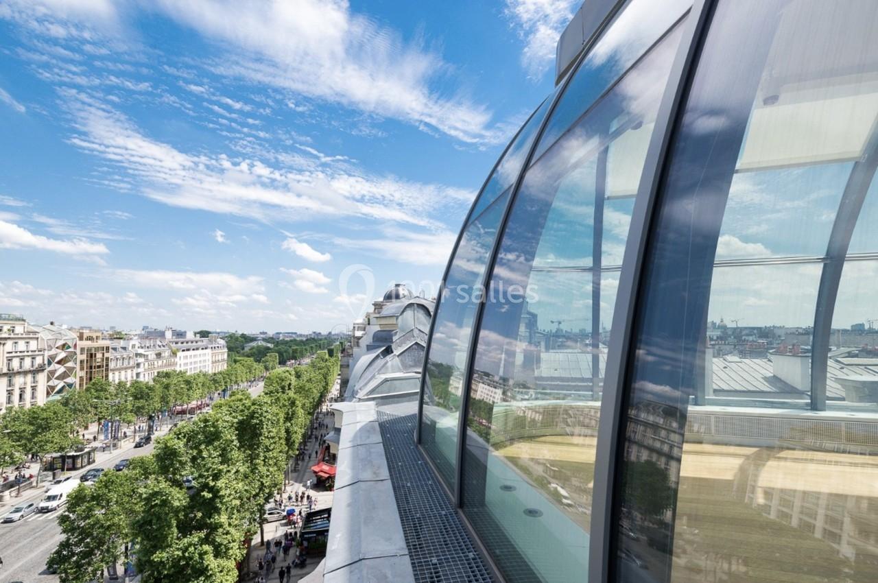 Vue d'une avenue bordée d'arbres et de bâtiments, reflétée dans une façade vitrée sous un ciel dégagé.