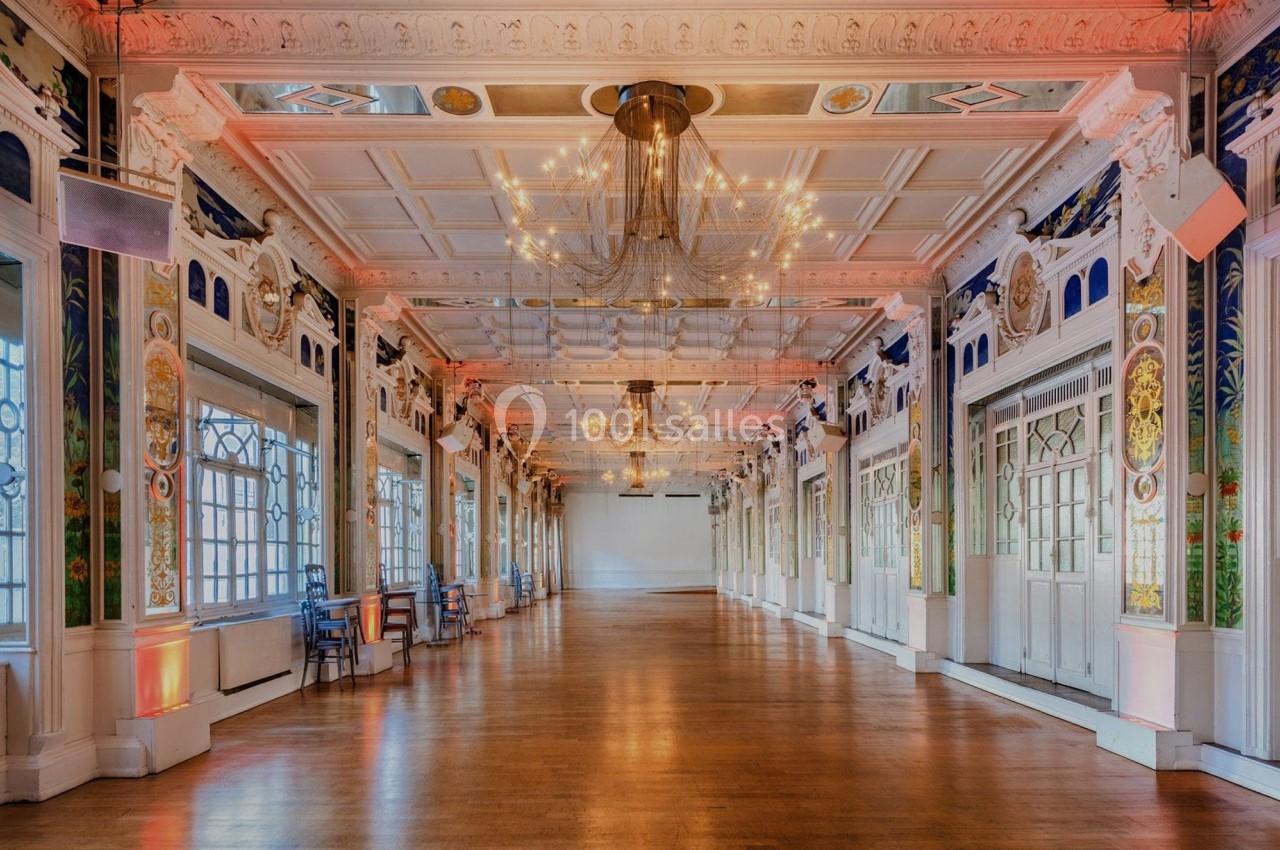 Salle spacieuse avec parquet, lustres suspendus et murs ornés de fresques et de grandes fenêtres.