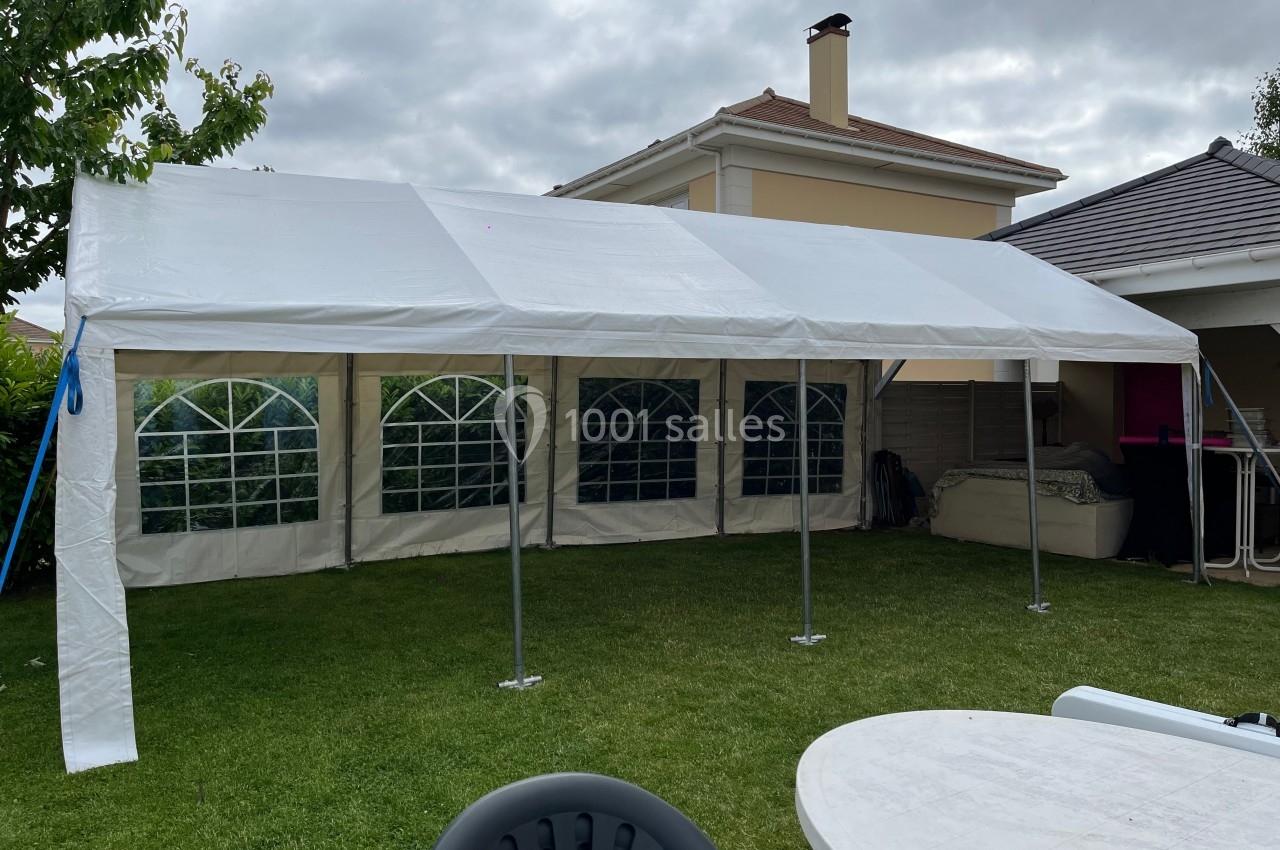 Tente blanche montée dans un jardin, avec des fenêtres en arc et des meubles de jardin visibles au premier plan.