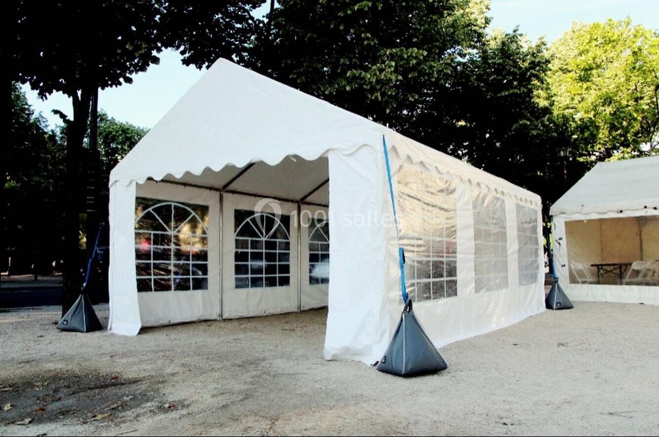 Tente blanche avec parois transparentes installée en extérieur sur un sol gravillonné, entourée d'arbres.