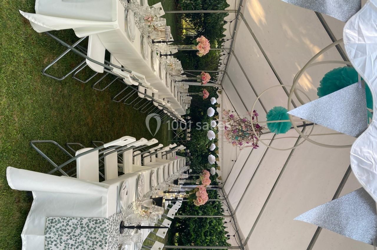 Tables dressées avec nappes blanches et décorations florales sous une tente en extérieur, prêtes pour un événement festif.