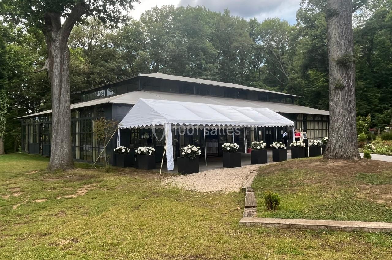 Bâtiment vitré entouré d'arbres avec une tente blanche à l'entrée et des jardinières de fleurs blanches.