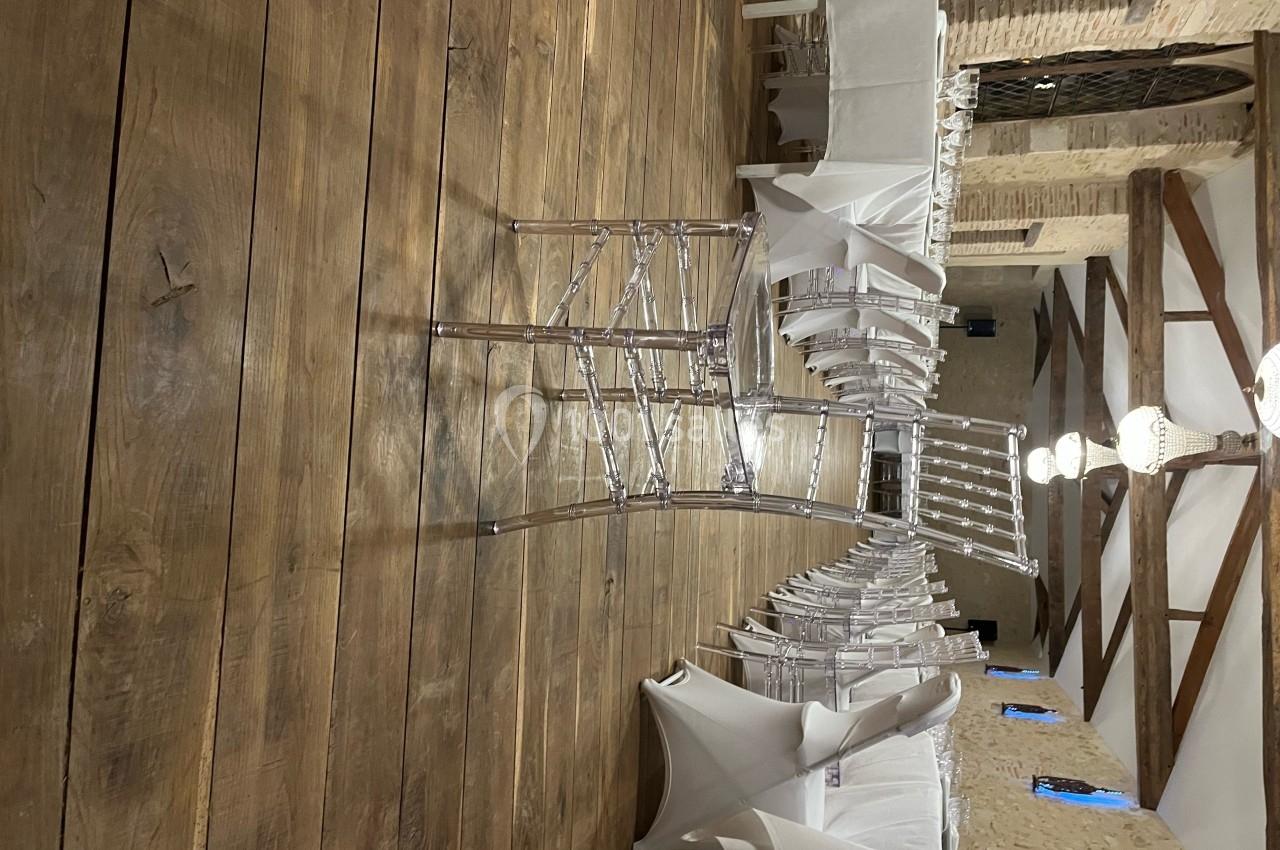 Salle de réception avec parquet en bois, chaises transparentes et nappes blanches, éclairée par des suspensions.