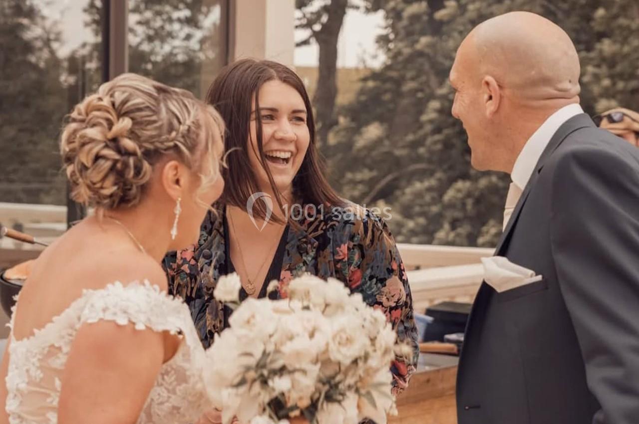 Une mariée, un homme en costume et une femme souriante discutent à l'extérieur lors d'une réception.