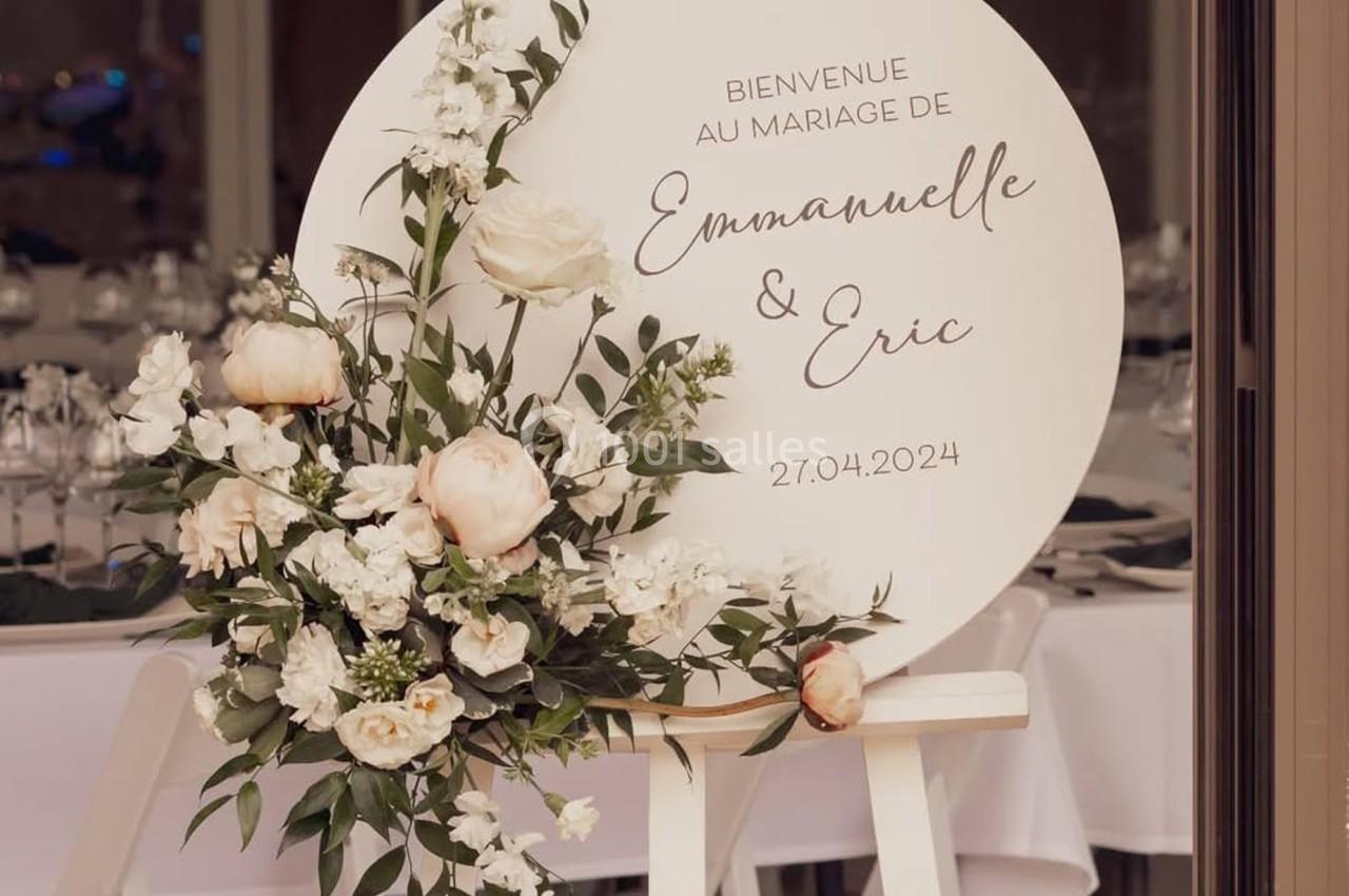 Panneau de mariage décoré de fleurs blanches et roses, avec les prénoms ’Emmanuelle & Eric’ et la date ’27.04.2024’.