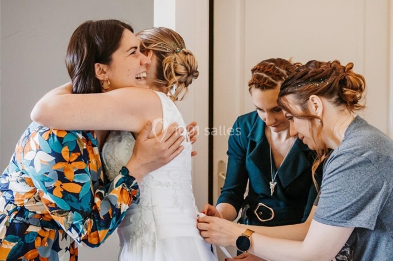 Deux femmes ajustent la robe d'une mariée pendant qu'une autre la serre dans ses bras, dans une ambiance joyeuse.