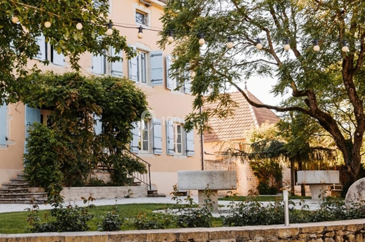 Façade d'une maison ancienne avec volets bleus, entourée de végétation et éclairée par des guirlandes lumineuses.