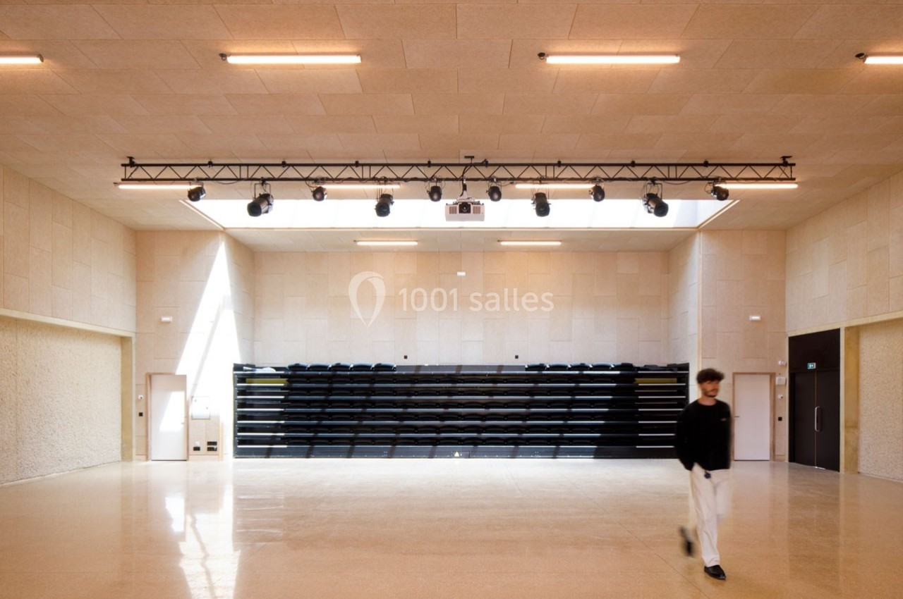 Salle lumineuse avec gradins repliés, éclairage suspendu et sol en béton poli, une personne marchant au premier plan.