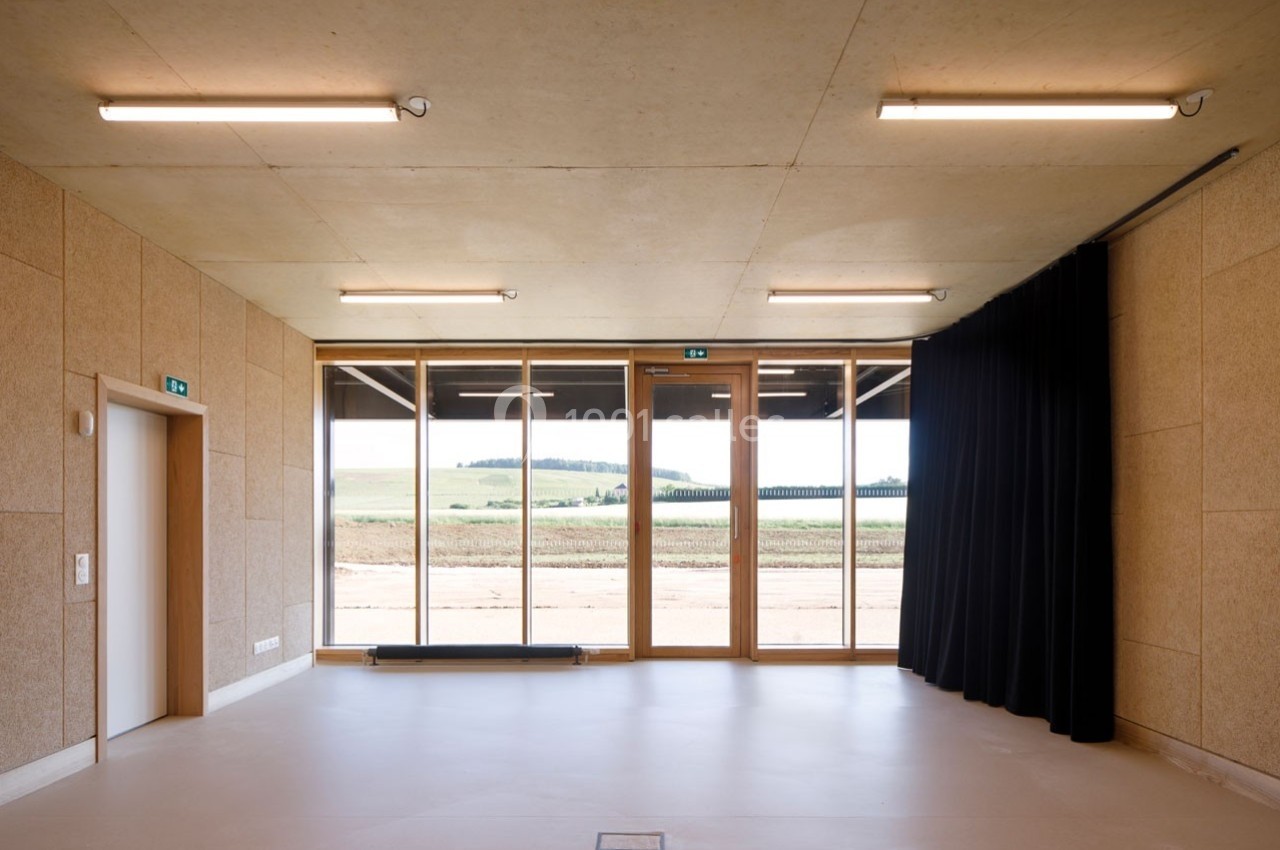 Salle lumineuse avec sol clair, murs en bois, grandes baies vitrées donnant sur un paysage champêtre et rideaux noirs.