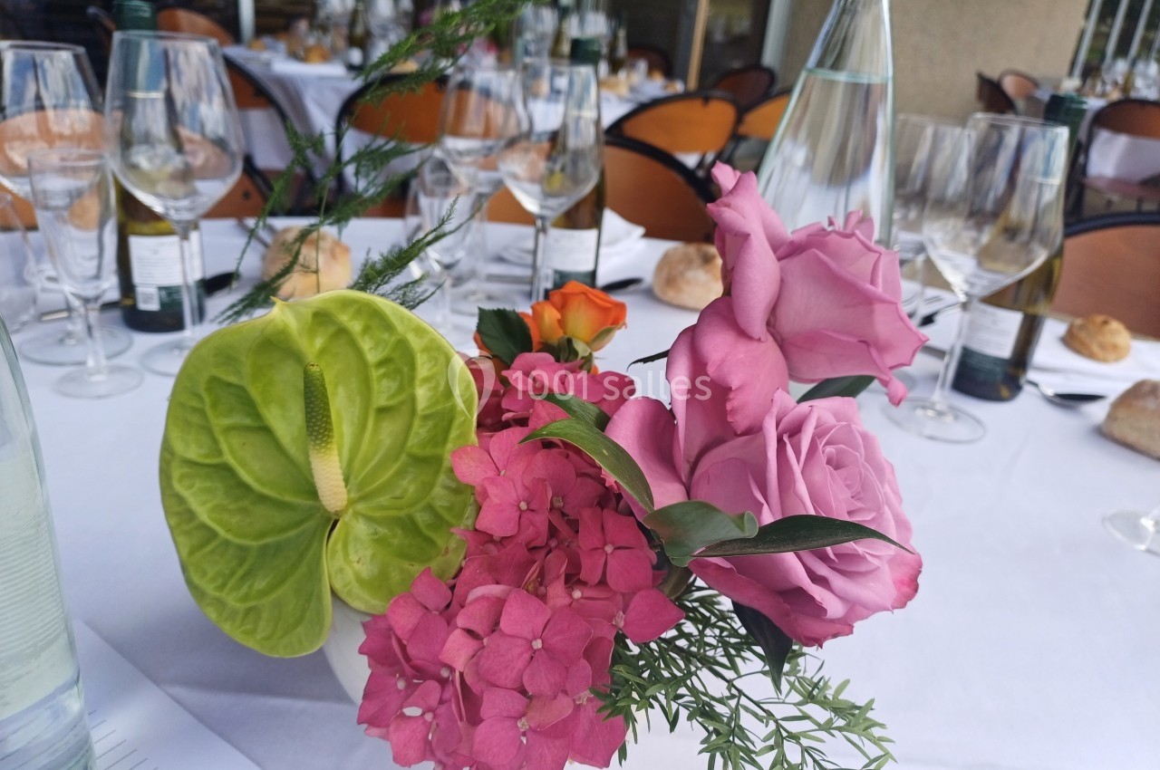Centre de table floral avec roses roses, hortensias, anthurium vert et feuillage, entouré de verres et bouteilles.