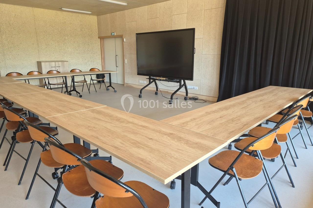 Salle de réunion avec tables disposées en U, chaises pliantes en bois et écran interactif sur pied.