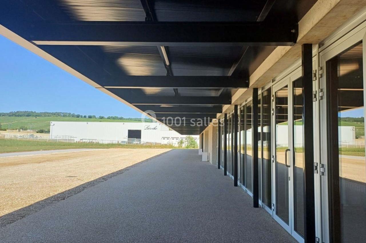 Allée couverte par une structure métallique noire, bordée de baies vitrées, avec un bâtiment industriel en arrière-plan.