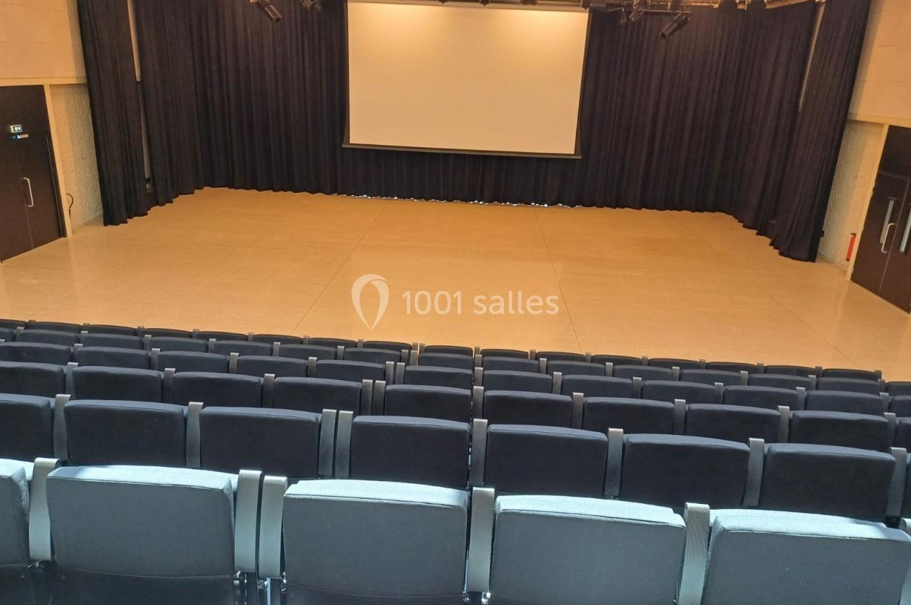 Salle de spectacle vide avec rangées de sièges noirs, scène en fond et écran blanc suspendu devant un rideau noir.