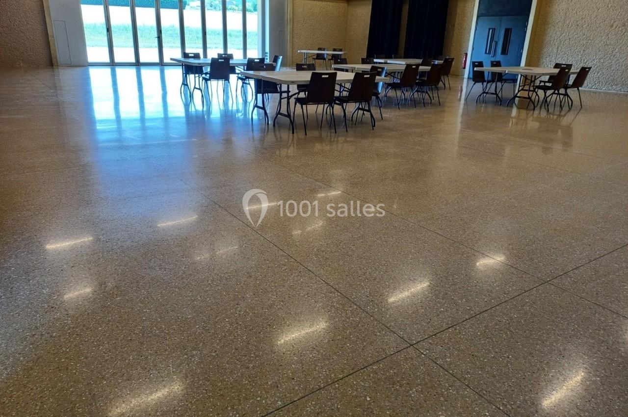 Salle spacieuse avec sol en terrazzo, tables et chaises disposées, grandes baies vitrées laissant entrer la lumière.