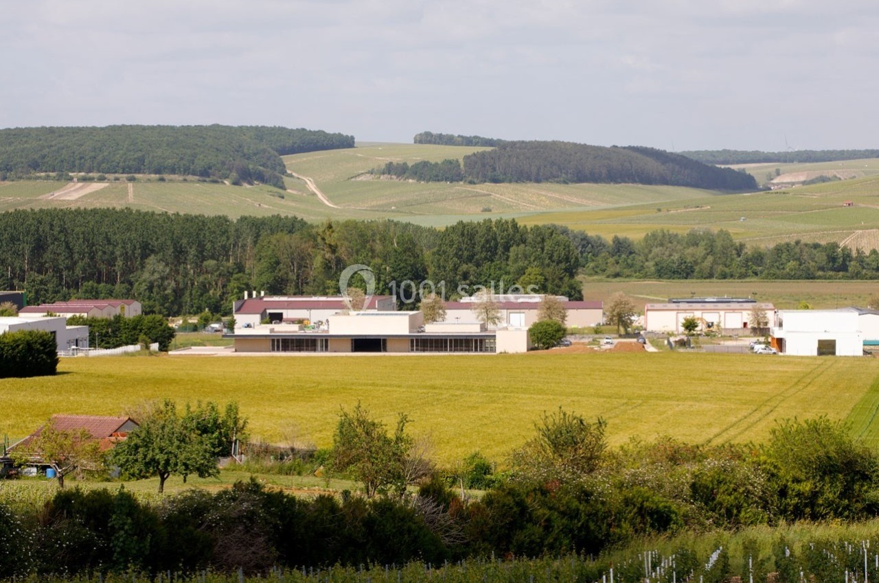 Paysage rural avec des champs, des bâtiments industriels et une forêt en arrière-plan sous un ciel partiellement nuageux.