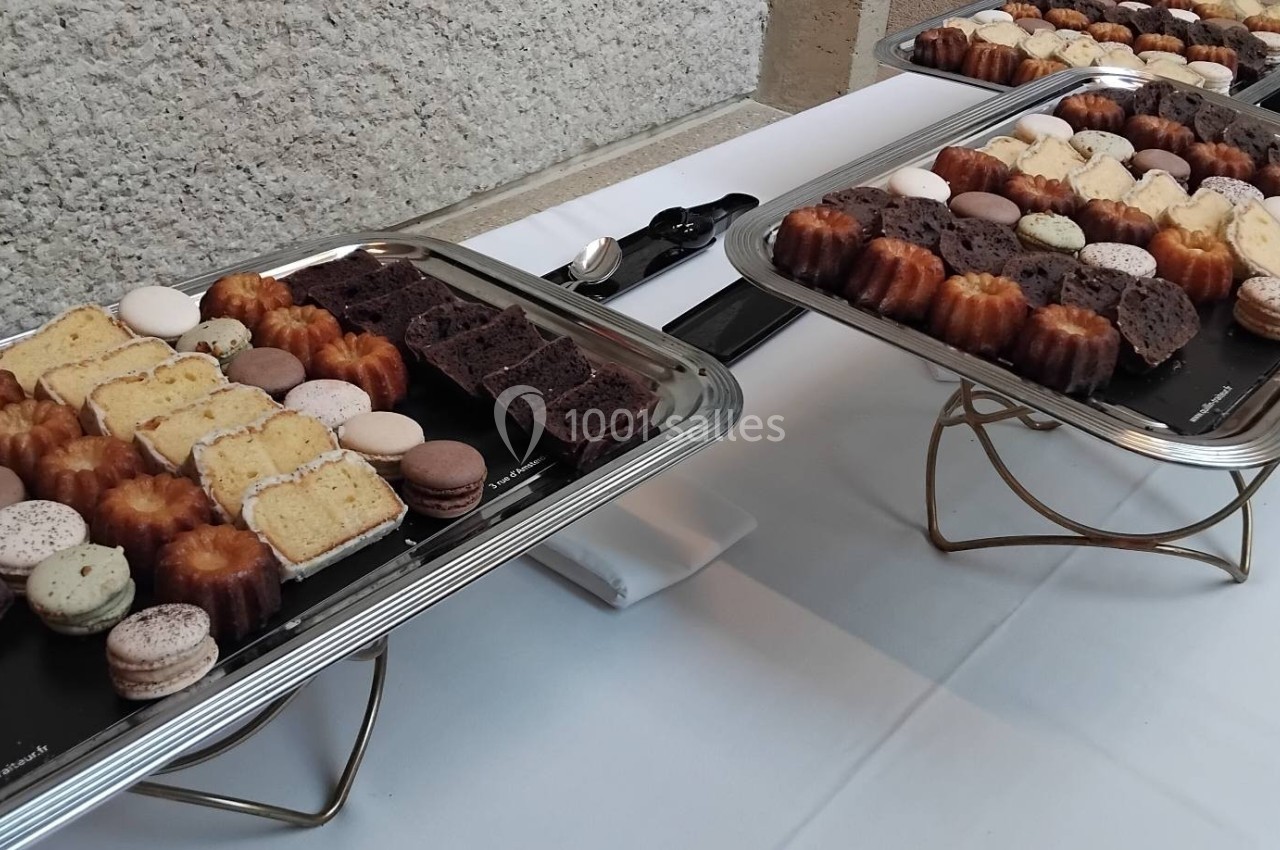 Plateaux de pâtisseries variées, incluant cannelés, macarons et tranches de cake, disposés sur une table blanche.