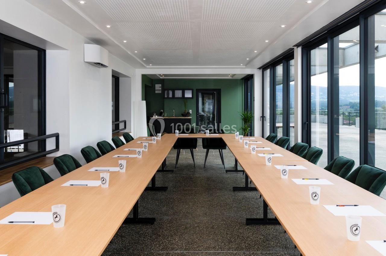 Salle de réunion lumineuse avec deux longues tables en U, chaises vertes, blocs-notes et vue sur l'extérieur.