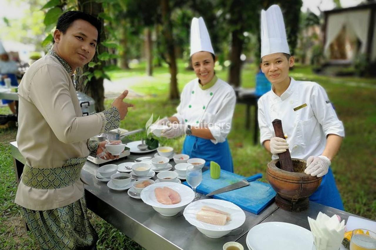 Trois cuisiniers en tenue professionnelle préparent des plats sur une table en extérieur, entourés de verdure.
