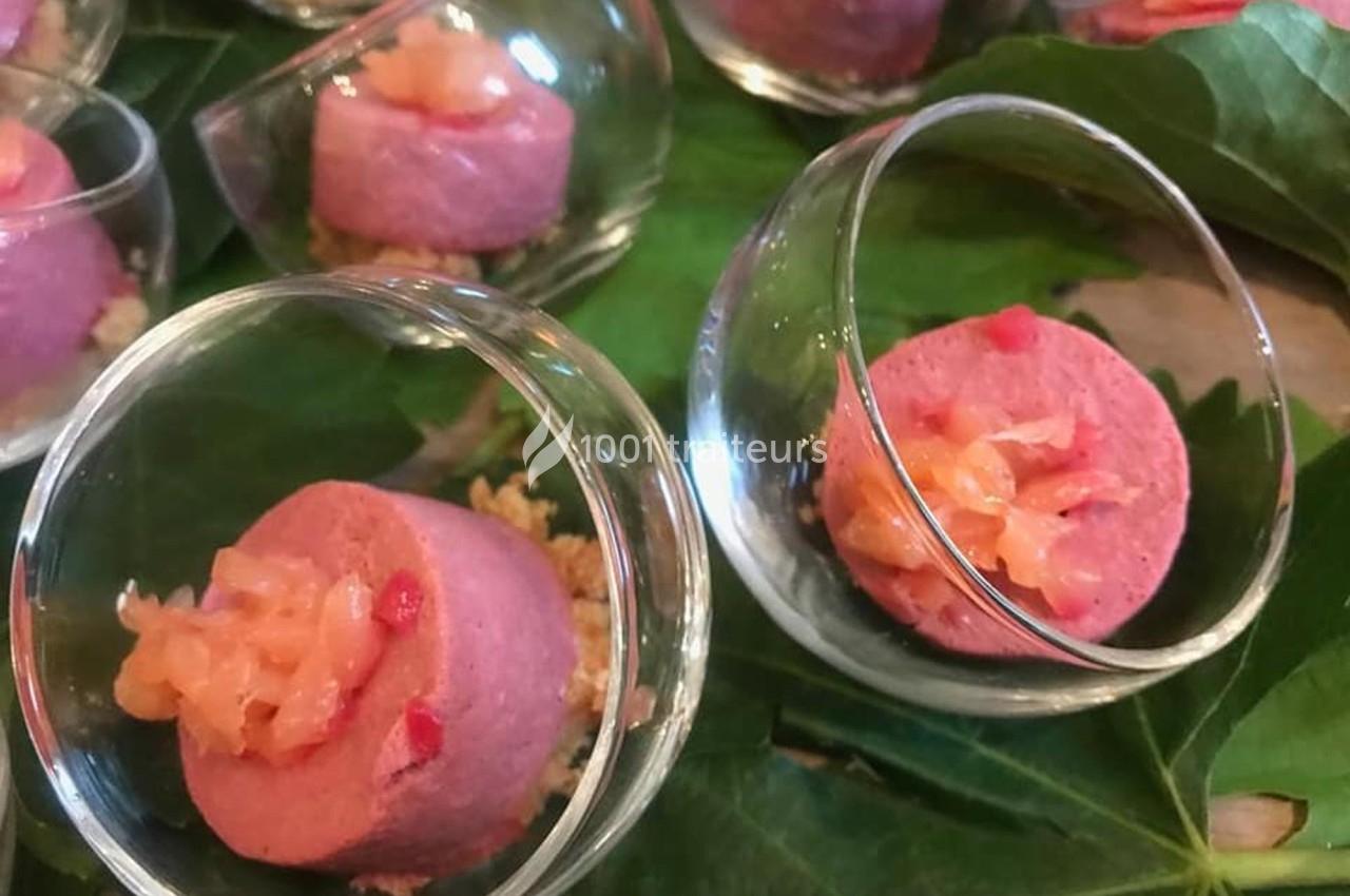 Verrines contenant une mousse rose garnie de morceaux de saumon sur un lit de crumble, présentées sur des feuilles vertes.