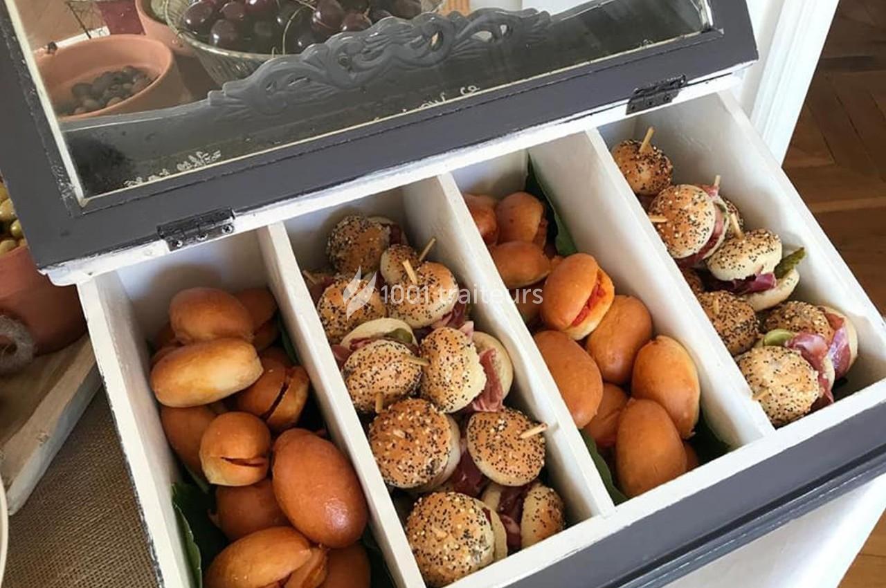 Plateau de mini-sandwichs variés disposés dans un coffret compartimenté, avec des fruits en arrière-plan.