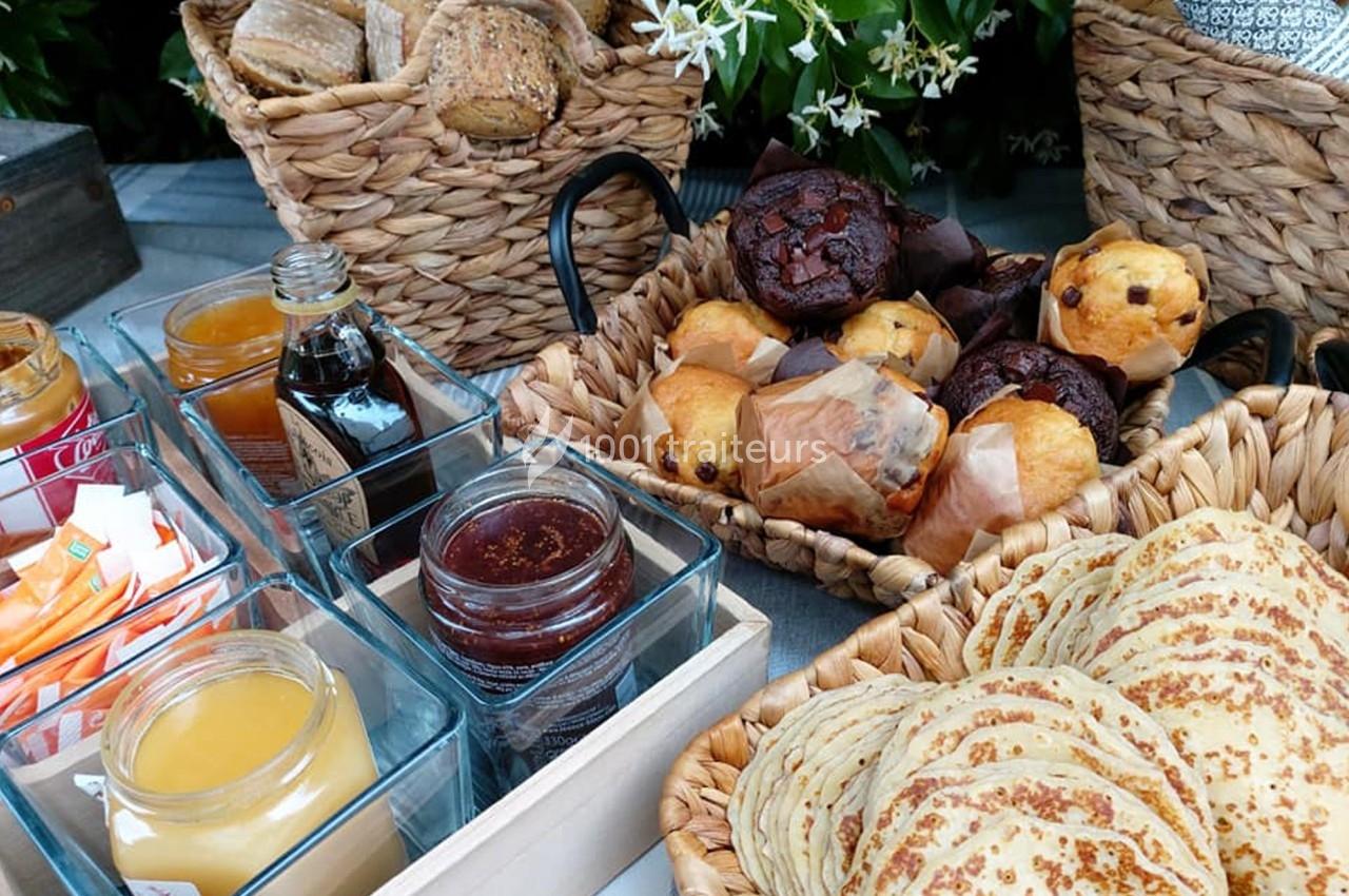 Assortiment de viennoiseries, crêpes, confitures et pains dans des paniers en osier sur une table extérieure.