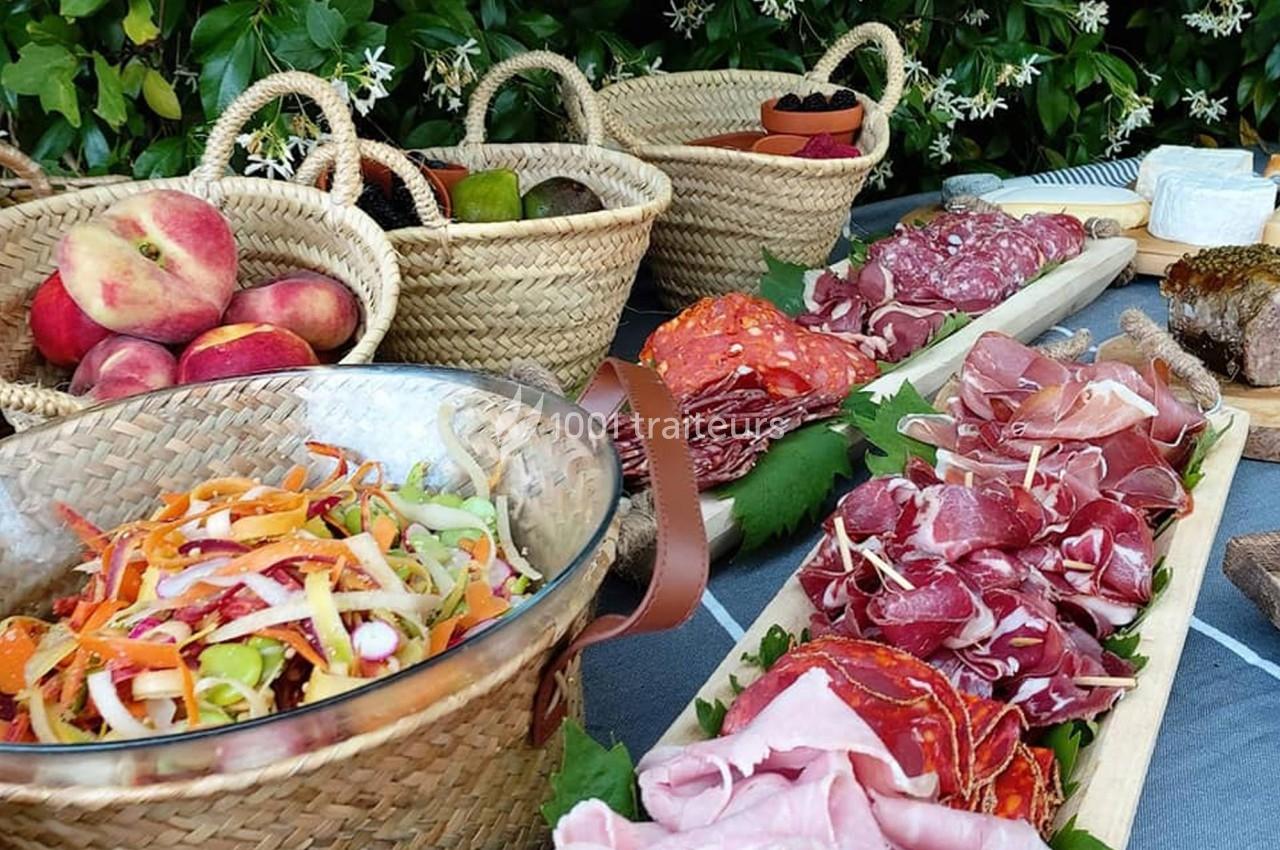 Plateau de charcuterie, salade colorée et paniers de fruits disposés sur une table en extérieur.